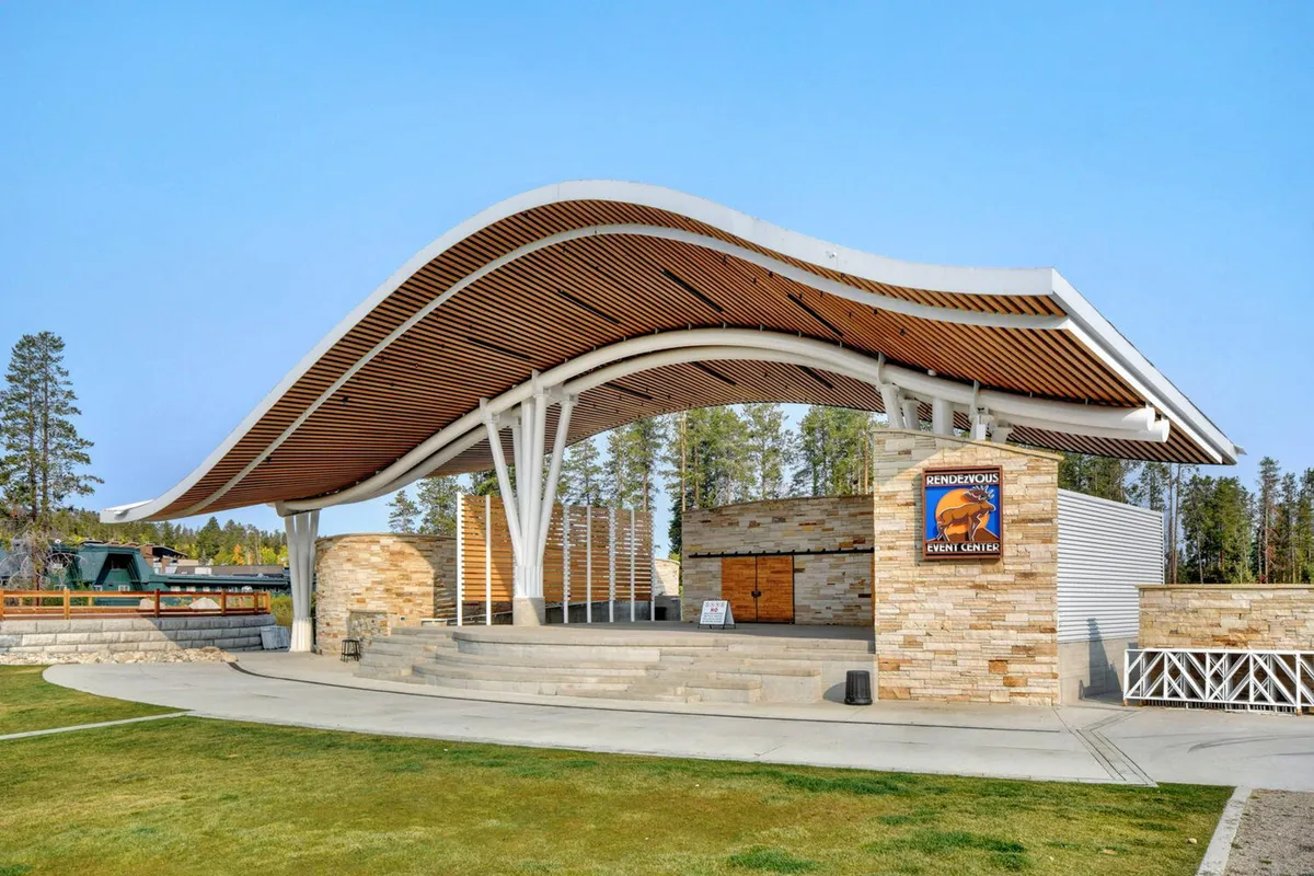 The image showcases the spacious steps and curved roof structure of the Hideaway Park Amphitheater, with people gathering for an outdoor concert or community event. The setting is open and communal, suggesting music performances and gatherings in the heart of the town.