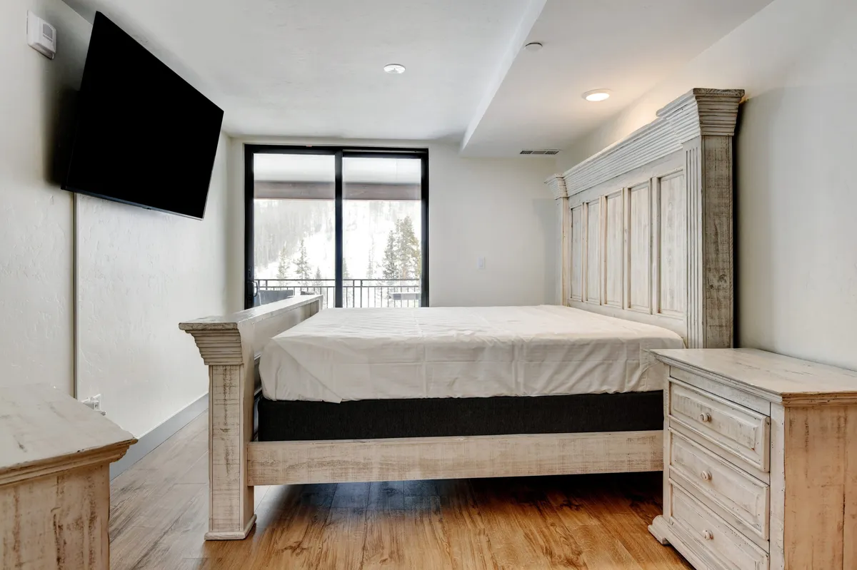 This image showcases a large bed with a rustic headboard, a flat-screen TV mounted on the wall, and large windows that let in abundant natural light with views of the outdoors. The overall vibe is calm and restful, inviting guests to relax or enjoy a peaceful evening watching TV or waking up to beautiful vistas.