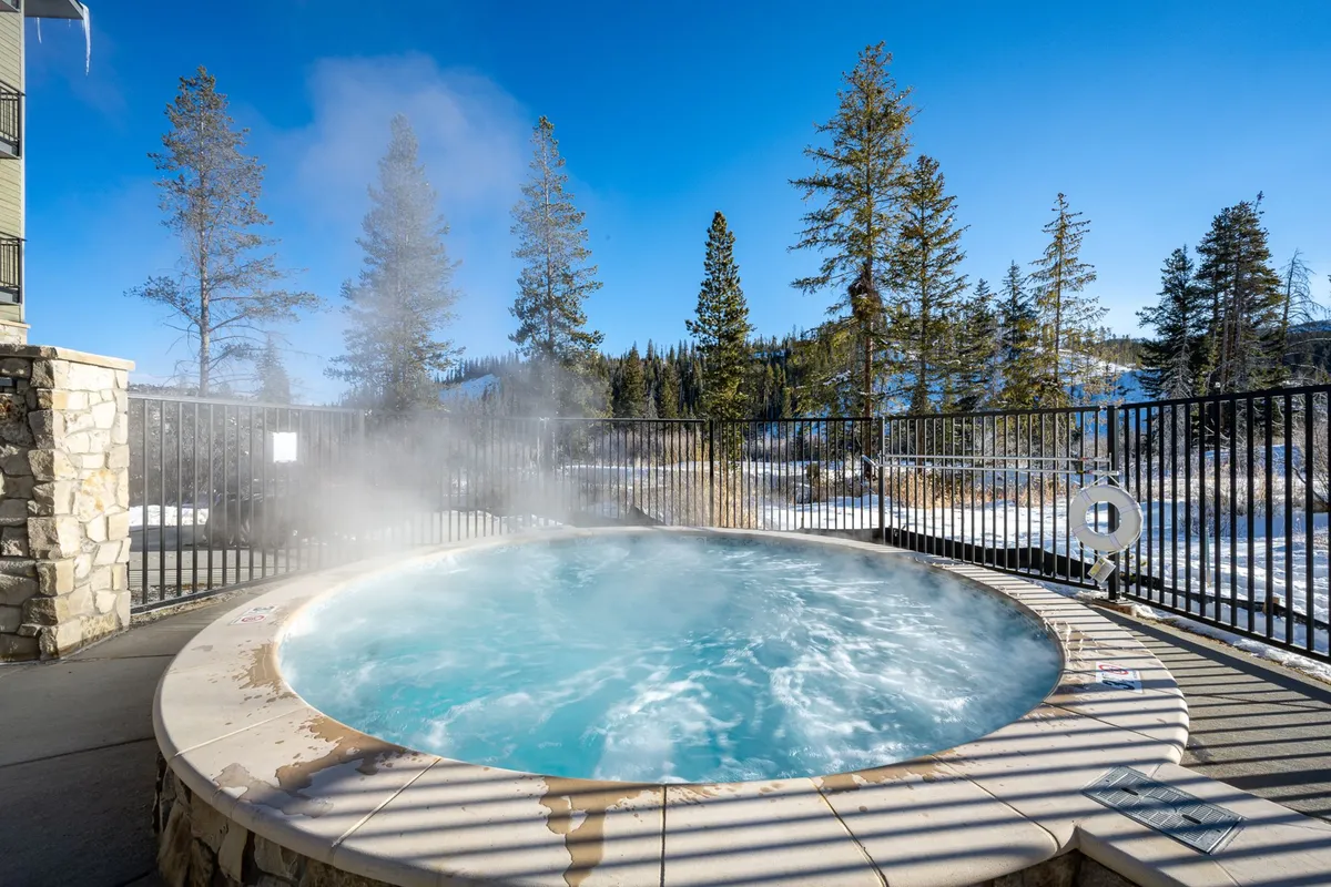 This exterior shot captures a bubbling hot tub surrounded by tall trees, with majestic mountain views and a clear blue sky overhead. The setting is tranquil and inviting, perfectly suited for evening relaxation or stargazing after a day of adventure.