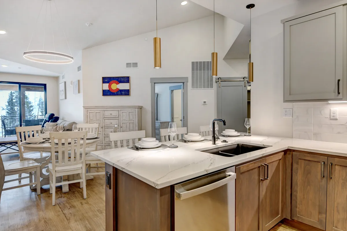 A bright, modern kitchen and dining area featuring a spacious kitchen island, tasteful artwork, and a dining table set for gathering. The open layout encourages socializing and the balcony hints at scenic views.