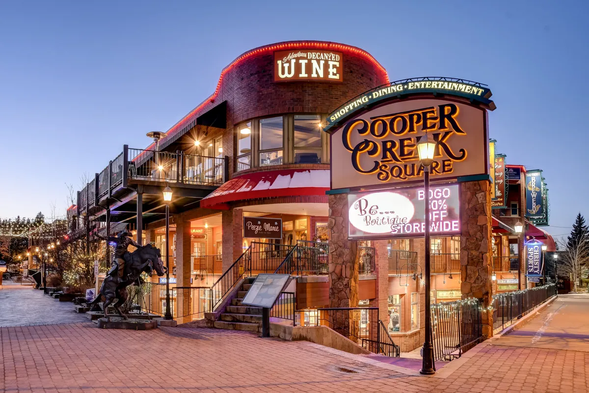 The scene features the Cooper Creek Square sign, a wine shop sign, an outdoor dining area filled with people, and a sculpture of a horse. The area is bustling with activity, evoking a lively atmosphere with shopping, dining, and entertainment in a pedestrian-friendly plaza.