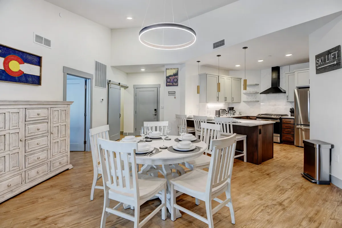 This inviting interior shot includes a stylish dining table set for five, a kitchen island in the background, a distinctive light fixture, and curated wall art. The mood is cozy and perfect for group meals or celebrations.