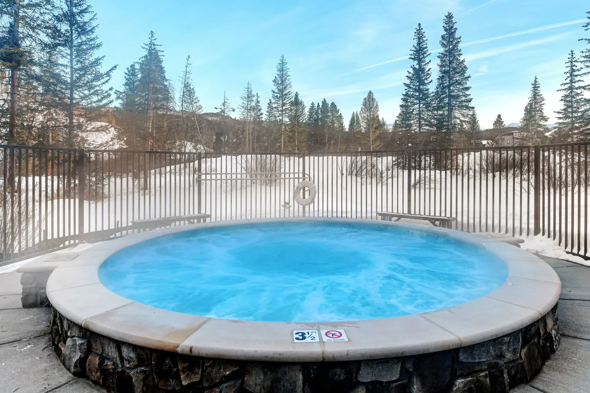 An outdoor hot tub sits amid a snowy landscape, encircled by pine trees. The scene evokes warmth and relaxation as steam rises from the tub, suggesting soothing soaks after skiing and opportunities for socializing under open skies.