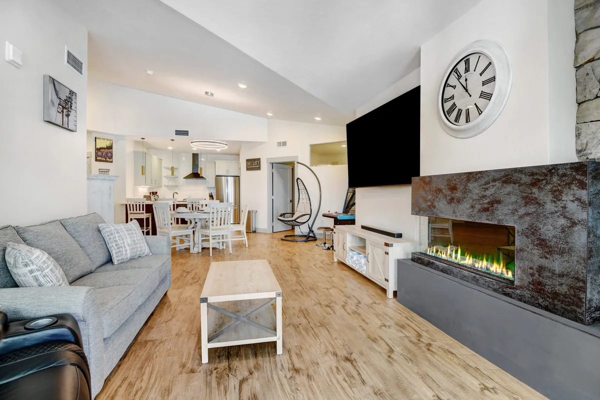 This interior image features a sophisticated open-plan living area with a plush sofa centered around a contemporary fireplace. The fireplace is the room's focal point, and an adjacent dining area is visible. Large windows flood the space with natural light and likely offer mountain views. The overall ambiance is cozy, modern, and inviting—perfect for group gatherings and relaxation.