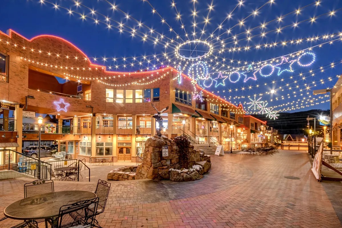 Twinkling lights are strung overhead against the evening sky, illuminating decorative star motifs and a central stone fountain. The lively outdoor plaza is surrounded by boutique shops and restaurants, inviting people to dine al fresco or stroll while enjoying community gatherings.
