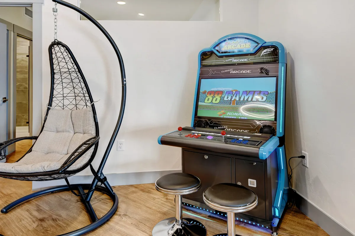 This interior image features a cozy nook with a hanging chair and a retro arcade game machine prominently displayed. The inviting space exudes a fun, nostalgic atmosphere, with the arcade cabinet as a focal point, perfect for both kids and adults looking to unwind. Soft lighting and relaxed seating make it ideal for socializing or solo play.