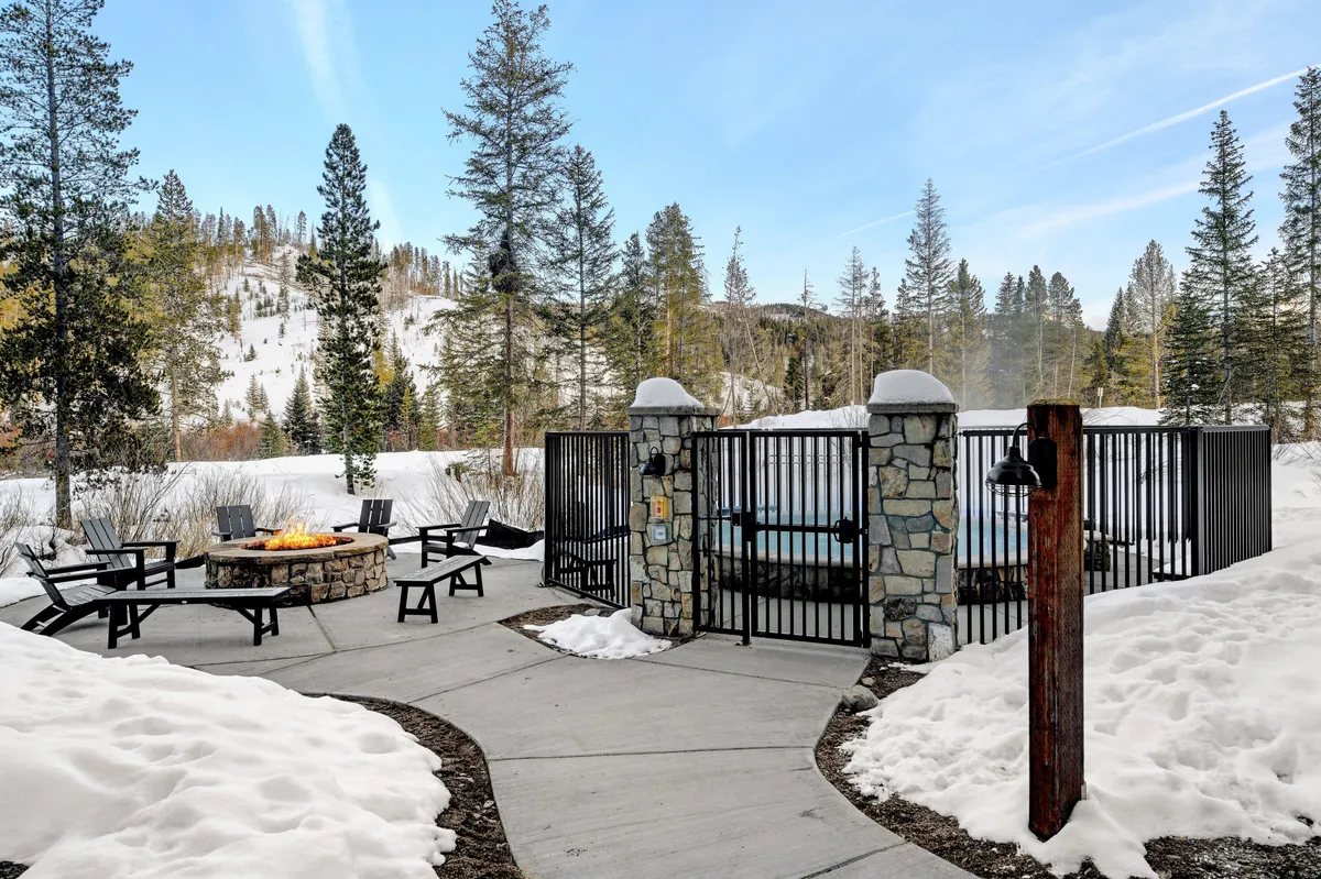 This image showcases a welcoming outdoor fire pit and an inviting hot tub, nestled among snow-covered ground and surrounded by tall trees. The setup encourages groups to gather around the fire or unwind in the hot tub while enjoying the peaceful natural setting.
