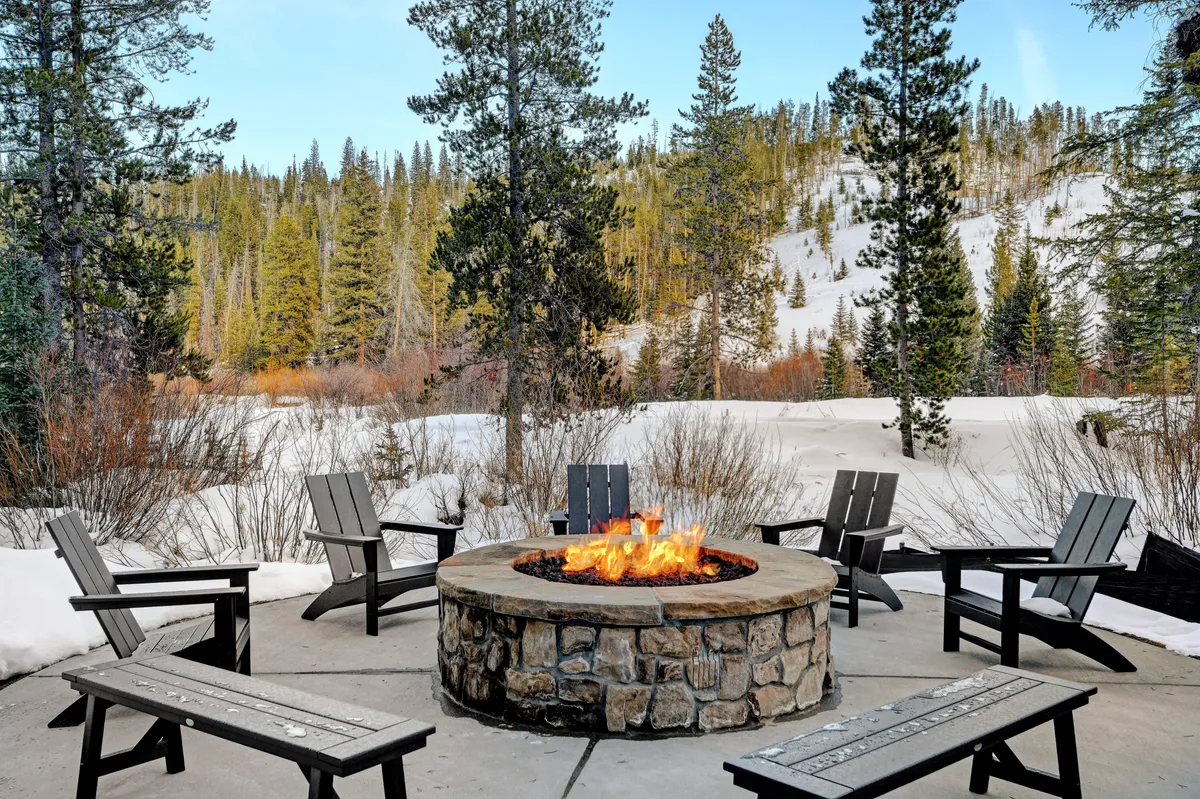 An inviting fire pit encircled by chairs on a snowy landscape, with distant trees and a cozy winter vibe. The setup encourages gathering for stories, warmth, and s’mores under the stars or at sunset.