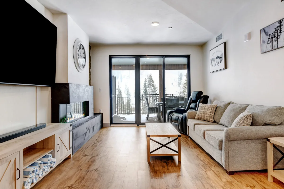 The image showcases a cozy living room with a plush sofa, modern coffee table, and a large TV, all framed by expansive windows that offer stunning sunrise views from the balcony. The inviting setup suggests comfort, warmth, and the perfect spot for relaxing or socializing after a day of adventure.